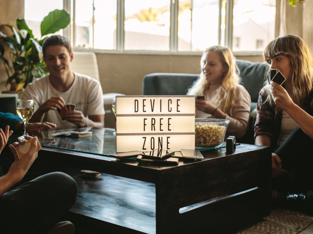 A group of friends enjoying a cozy game night around a table, laughing and playing cards. A glowing sign reads "DEVICE FREE ZONE", highlighting the joy of screen-free fun. This moment captures the magic of bringing people together for real connection and laughter.