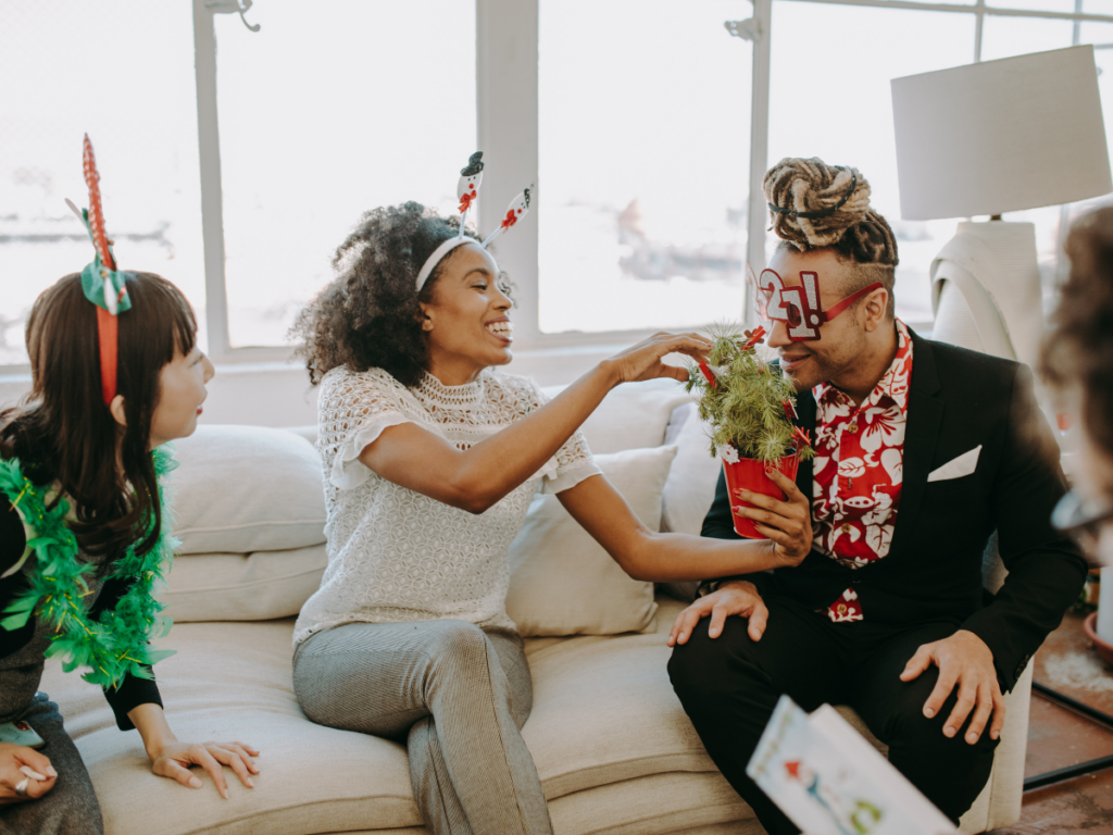 A group of friends enjoying a festive gathering, wearing holiday accessories and laughing while playing a fun game. One person playfully hands a small decorated plant to another, creating a joyful and interactive moment. This scene highlights upcoming events where printable games bring people together for laughter and connection.