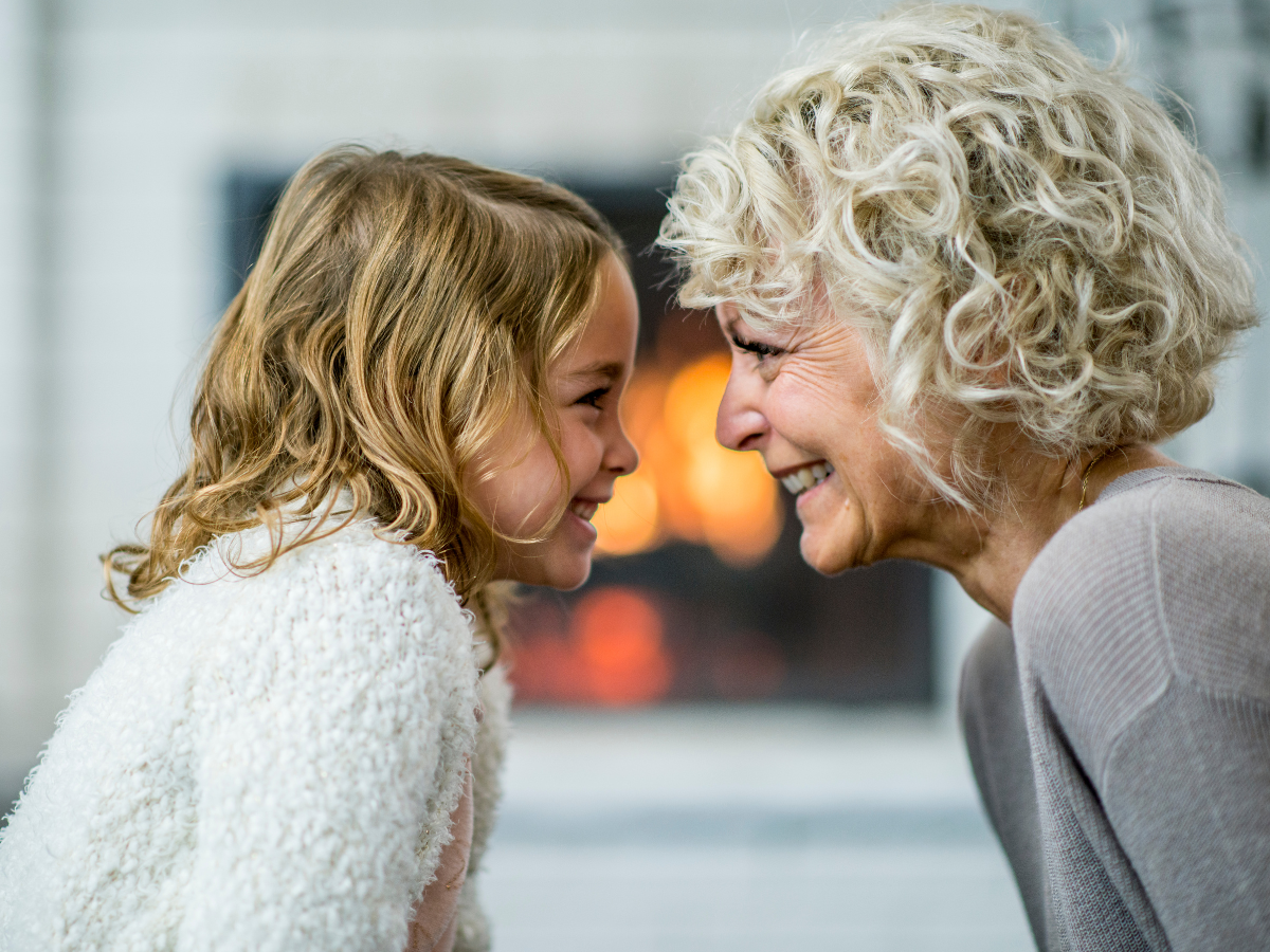 Happy grandmother and young girl smiling face-to-face by the fireplace, showing emotional connection and the benefits of face-to-face communication over screens with printable games.