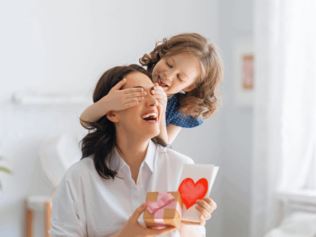A smiling mother being surprised by her daughter with a handmade card and wrapped gift, capturing the joy of Mother’s Day.