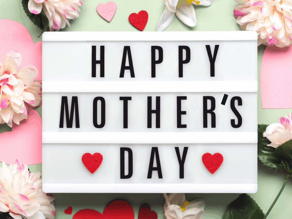Lightbox sign displaying “Happy Mother’s Day” surrounded by pink paper hearts and fresh spring flowers for a festive touch.